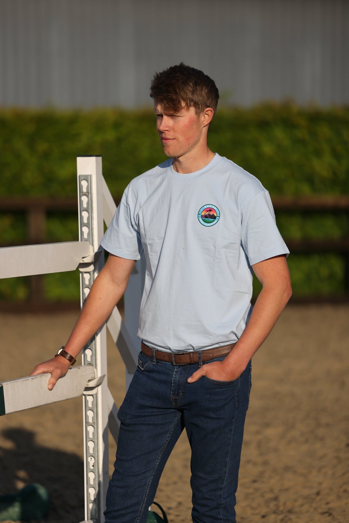 A young man standing in an outdoor equestrian arena wearing a light blue crewneck t-shirt with a circular logo on the left chest, paired with dark blue jeans.