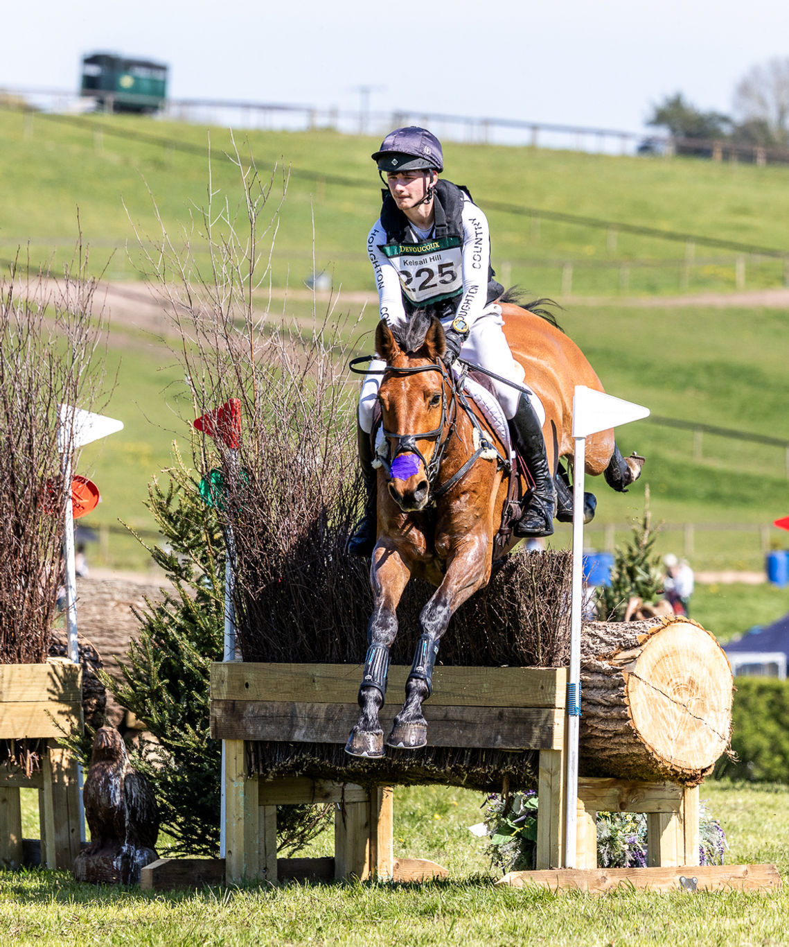 A horse rider on a brown horse jumps a wooden log obstacle in an outdoor equestrian event, with grassy hills and trees in the background.
