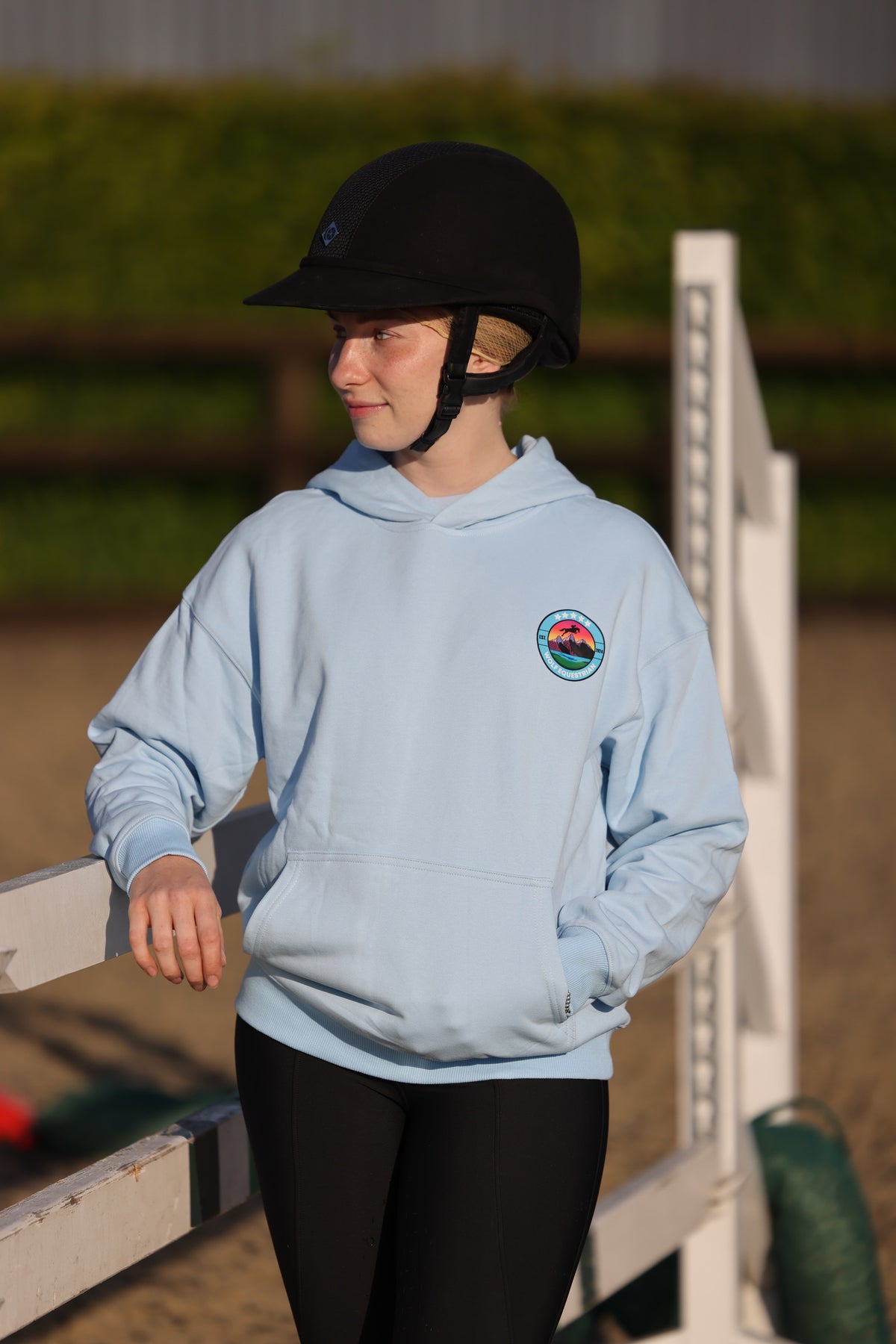 A child wearing a light blue equestrian hoodie with a circular crest on the left chest, black riding breeches, and a black riding helmet, standing in an outdoor equestrian arena.