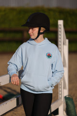 A child wearing a light blue equestrian hoodie with a circular crest on the left chest, black riding breeches, and a black riding helmet, standing in an outdoor equestrian arena.