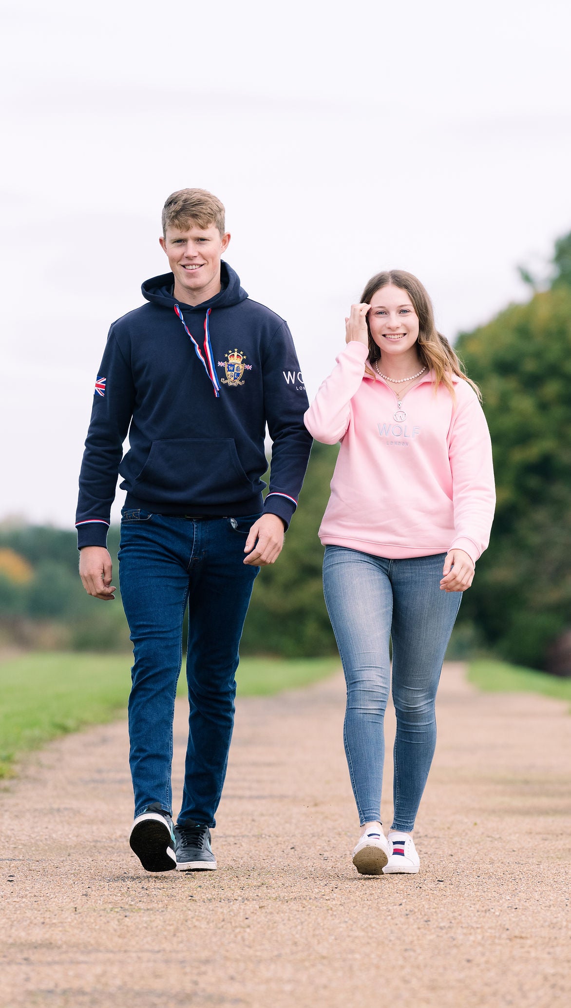 A young man and woman walking side-by-side on a gravel path outdoors. The man is wearing a navy blue hoodie with a crest on the left chest and 'WEL' on the sleeve, paired with dark blue jeans.