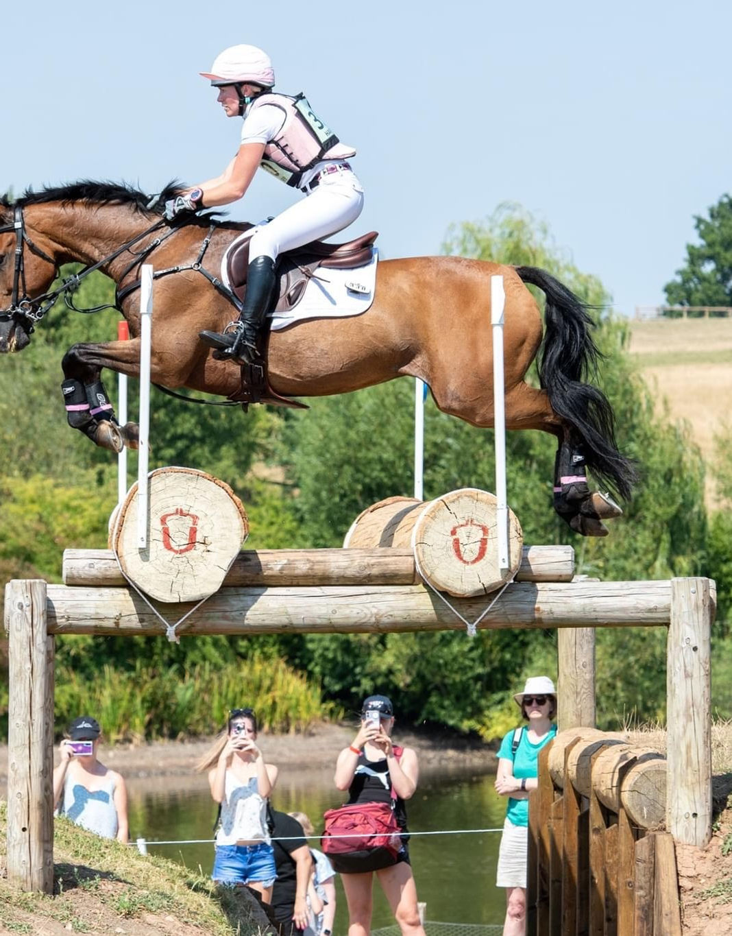 A horse rider on a brown horse jumps a wooden obstacle over water during an equestrian event, with spectators watching from the sidelines.