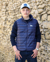A young man wearing a navy blue puffer vest with a white embroidered bear logo, a dark blue ribbed sweater, dark trousers, and a white cap, standing in front of a rustic stone wall.