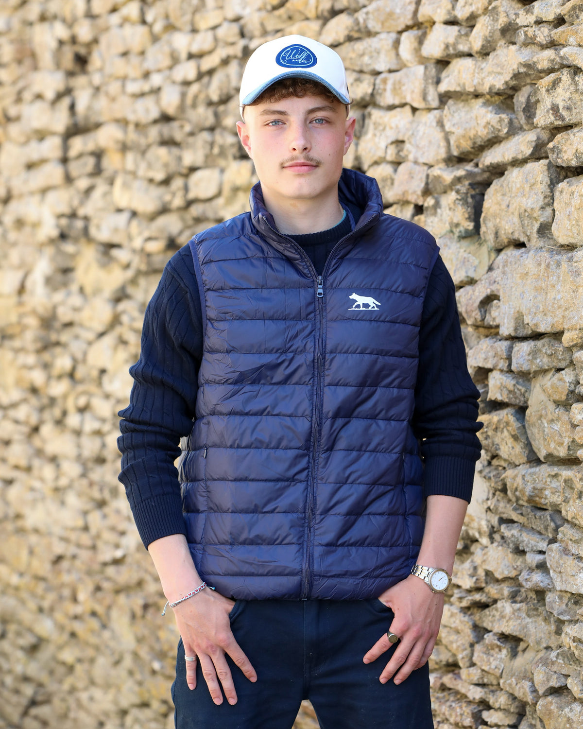A young man wearing a navy blue puffer vest with a white embroidered bear logo, a dark blue ribbed sweater, dark trousers, and a white cap, standing in front of a rustic stone wall.