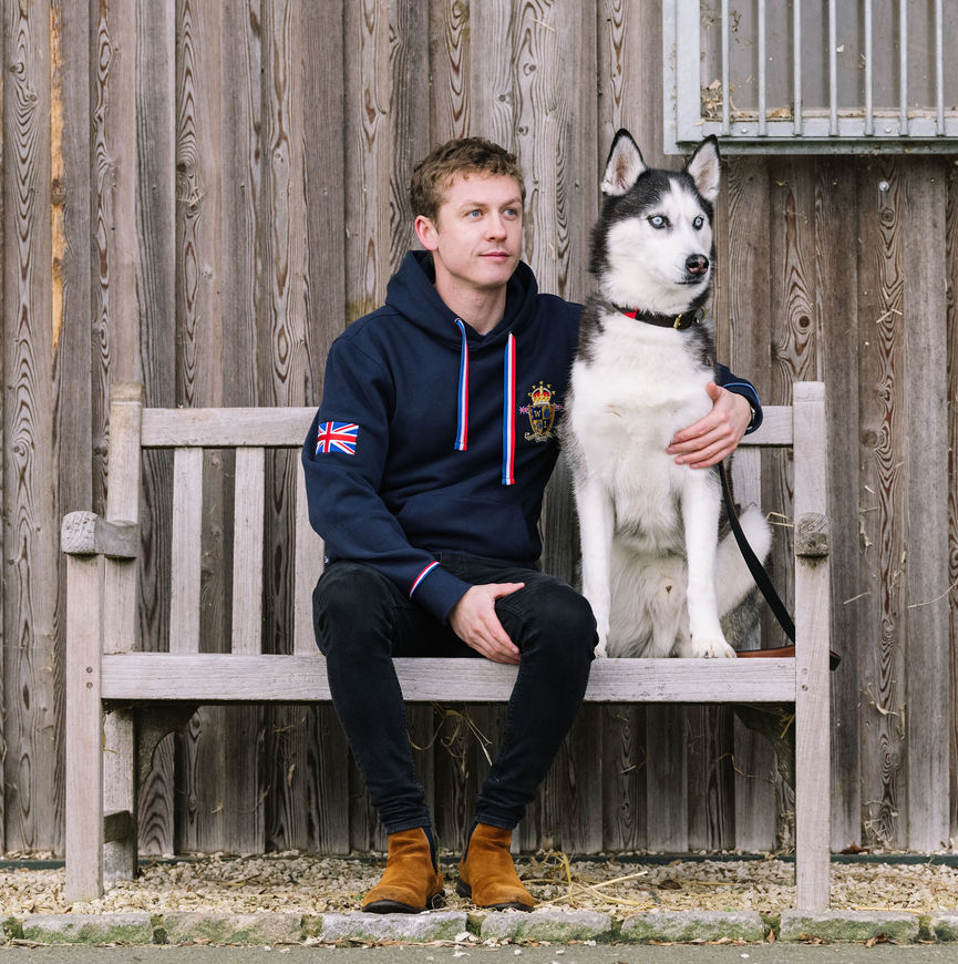 Andre Founder wearing a navy blue hoodie with a Union Jack patch on the sleeve sits on a wooden bench next to Storm a husky dog. The background is a weathered wooden fence.