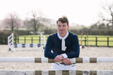 A young man in equestrian attire, wearing a navy blue long-sleeve shirt, white collar, and white breeches, leaning on a jumping fence in an outdoor equestrian arena.