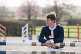 A young man in equestrian attire, wearing a navy blue sweater, white shirt, and white tie, leaning on a jumping fence at an outdoor equestrian arena.