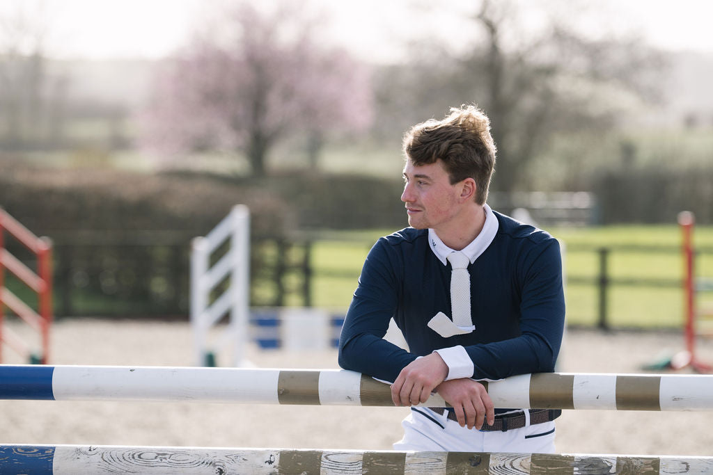 A young man in equestrian attire, wearing a navy blue sweater, white shirt, and white tie, leaning on a jumping fence at an outdoor equestrian arena.
