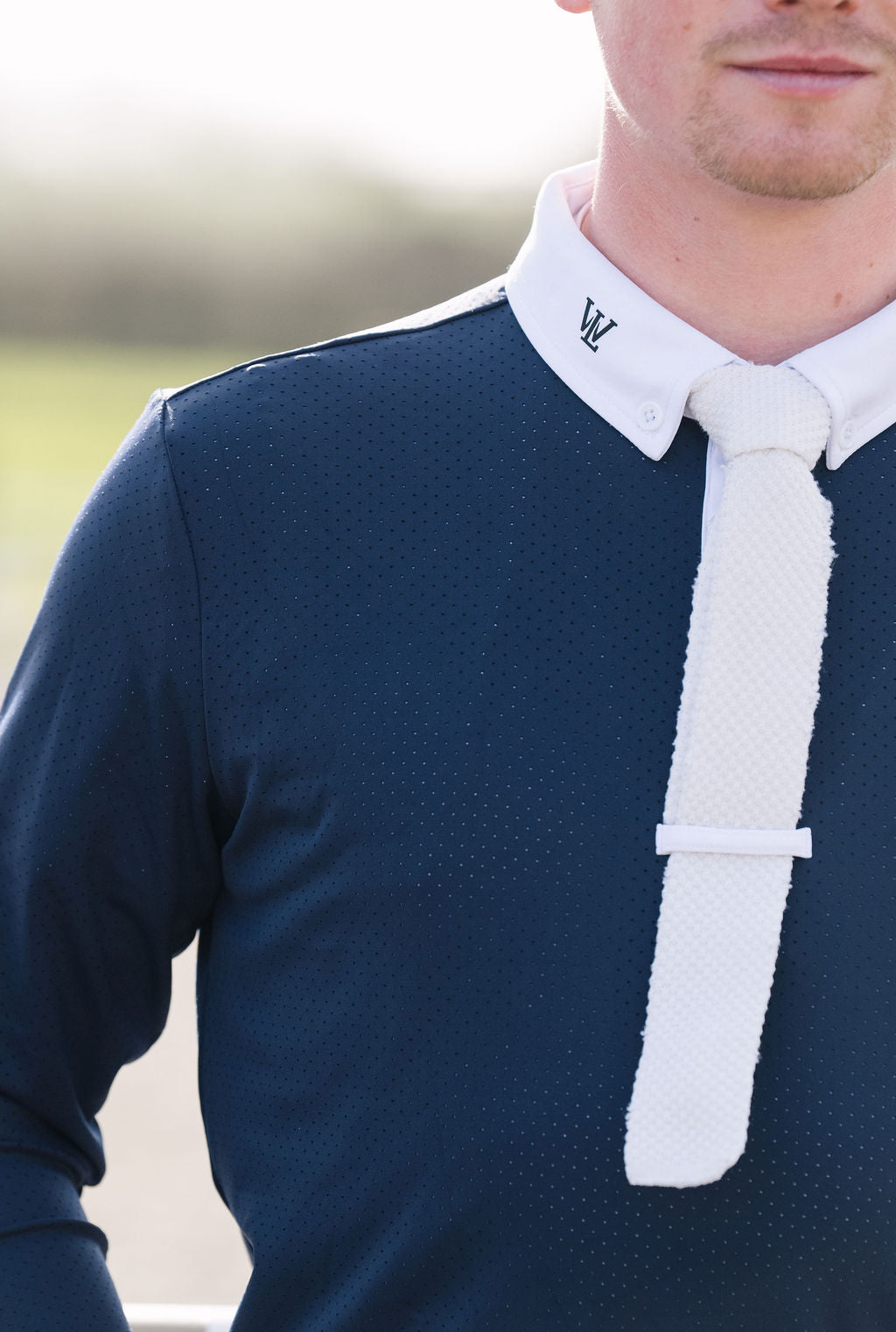 Close-up of a man wearing a navy blue textured long-sleeve shirt with a white collar featuring a black embroidered monogram 'W' and a matching white tie.