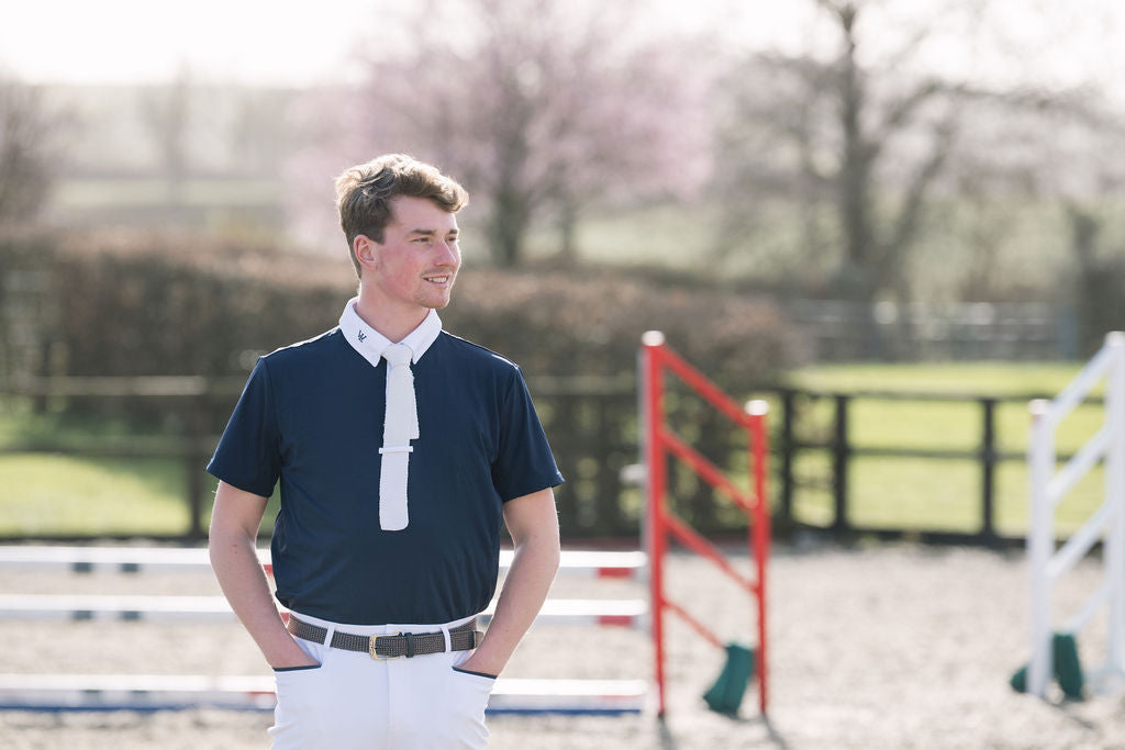 A young man standing outdoors in an equestrian arena, wearing a navy men's competition shirt with a white collar and white trousers.