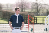 A young man standing outdoors in an equestrian arena, wearing a navy men's competition shirt with a white collar and white trousers.