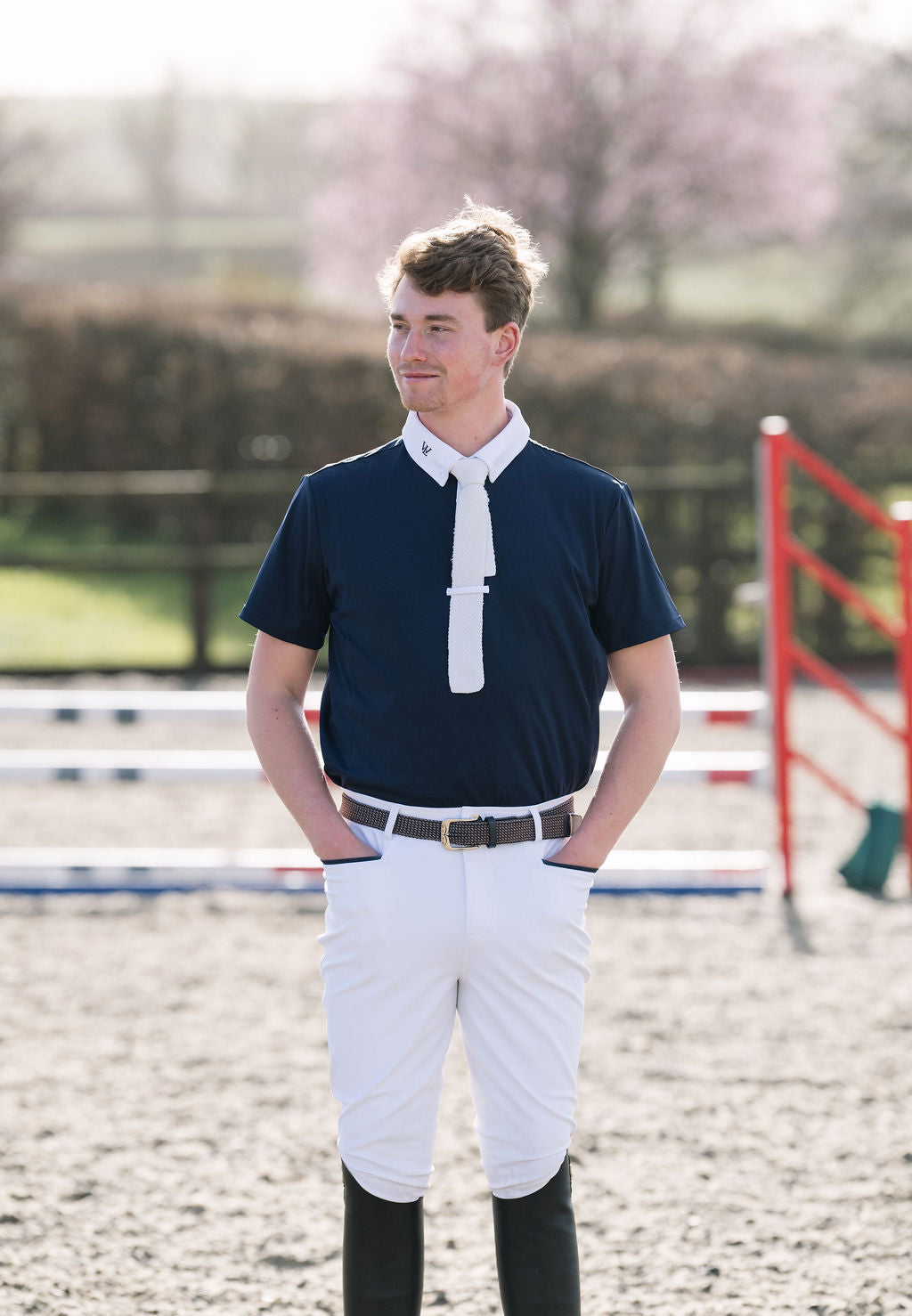 A young man standing in an equestrian arena wearing a navy men's competition  shirt with a white necktie detail, white breeches, and black riding boots.