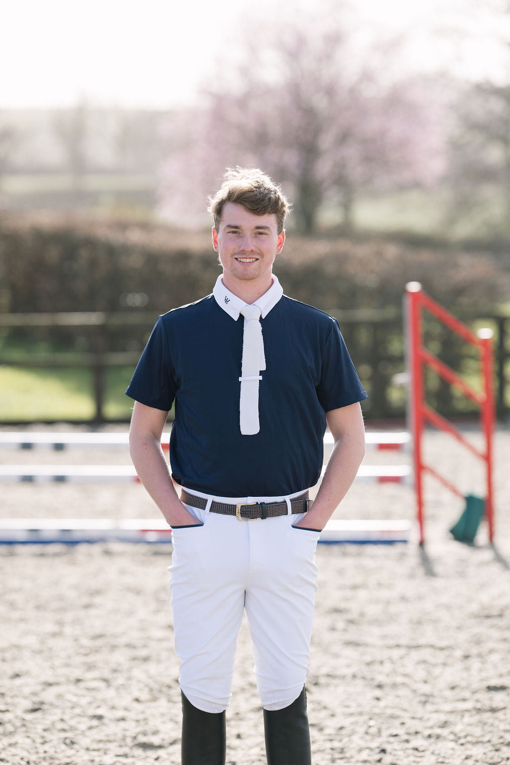 A man wearing a navy men's competition  shirt with a white collar and white tie, white riding breeches, and black riding boots, standing in an equestrian arena.