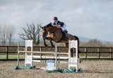 A horse rider in a navy men's competition Shirt and white pants jumps a horse over an outdoor show jumping obstacle in an equestrian arena.