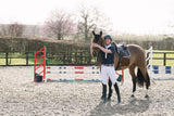 A person in Wolf London equestrian attire stands with a brown horse in an outdoor riding arena, with a jump and trees in the background.