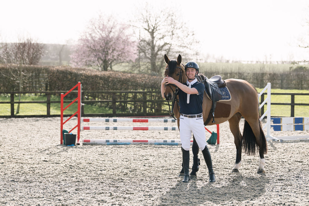 A person in Wolf London equestrian attire stands with a brown horse in an outdoor riding arena, with a jump and trees in the background.