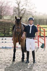A man in equestrian attire stands beside a brown horse in an outdoor riding arena. He is wearing a navy men's competition shirt, white breeches, black riding boots, and a dark helmet.