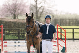 A man in equestrian attire, wearing a navy helmet, navy wolf equestrian polo competition shirt white tie and white trousers, standing next to a brown horse in an outdoor riding arena with jump rails and trees in the background.