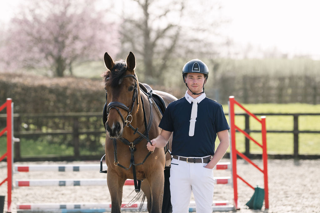 A man in equestrian attire, wearing a navy helmet, navy wolf equestrian polo competition shirt white tie and white trousers, standing next to a brown horse in an outdoor riding arena with jump rails and trees in the background.