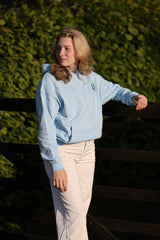 A woman wearing a light blue hoodie and cream pants, leaning against a dark wooden fence with a green hedge background.