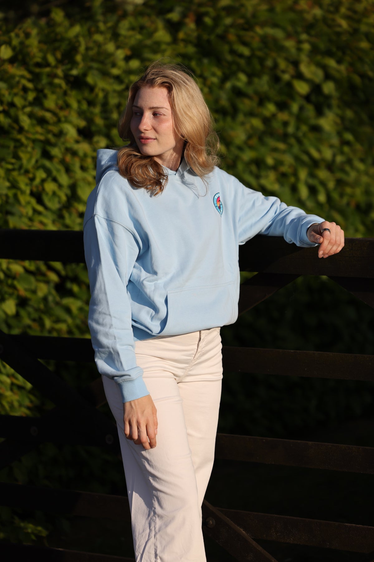 A woman wearing a light blue hoodie and cream pants, leaning against a dark wooden fence with a green hedge background.