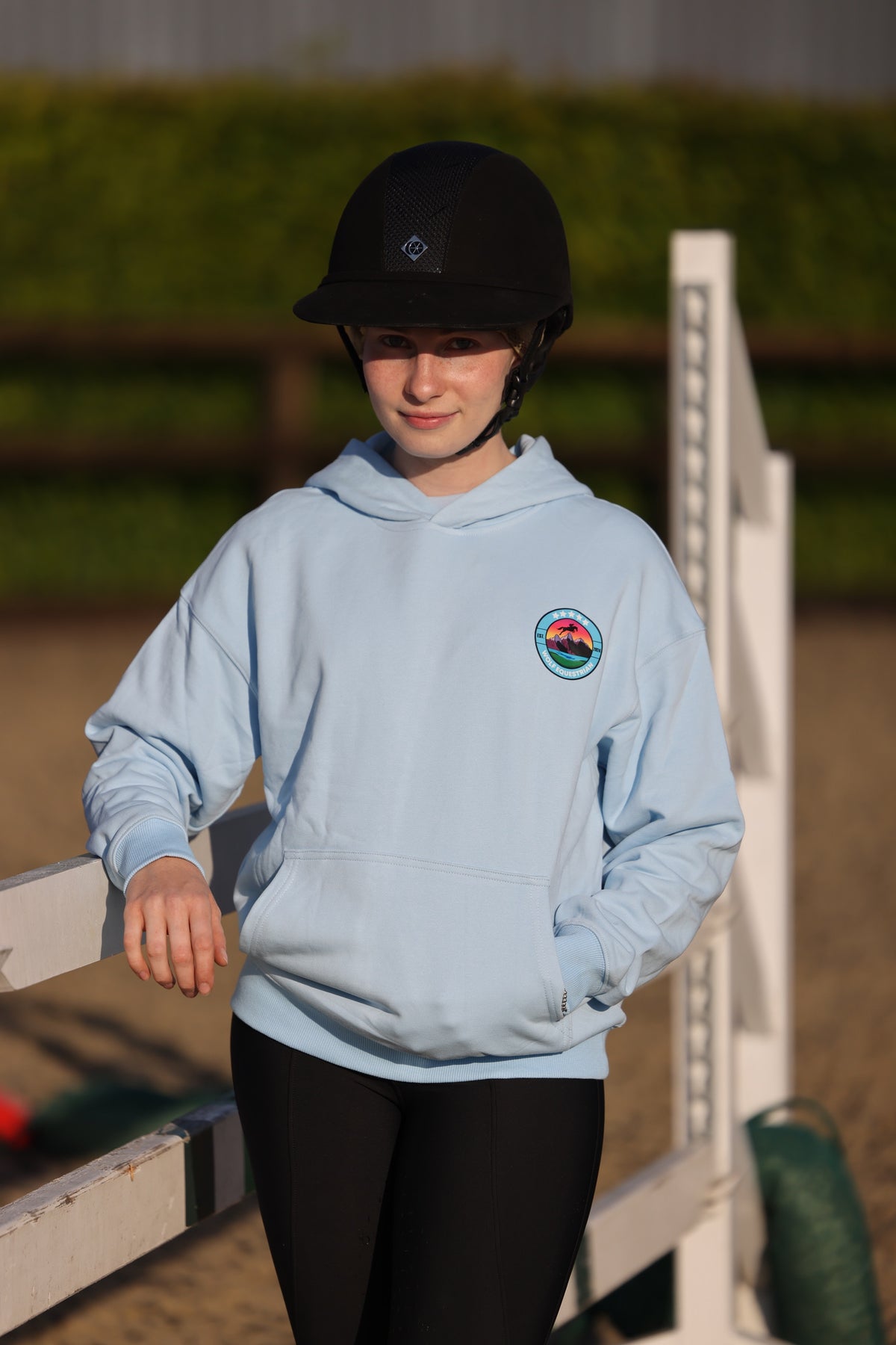 A person wearing a light blue hoodie with a circular equestrian crest on the left chest, black riding breeches, and a black riding helmet, standing outdoors near a white jump rail in an equestrian arena.