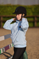 A young rider wearing a black equestrian helmet and a light blue hoodie with a circular logo on the chest, standing in an outdoor equestrian arena.