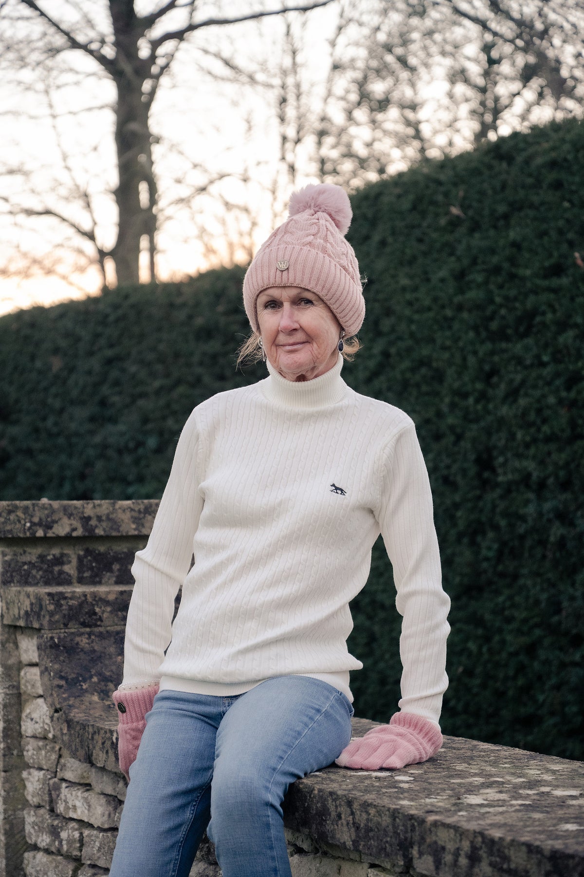 A woman wearing a cream ribbed turtleneck sweater, a light pink knit beanie with a pom-pom, and pink gloves, sitting on a stone wall outdoors.