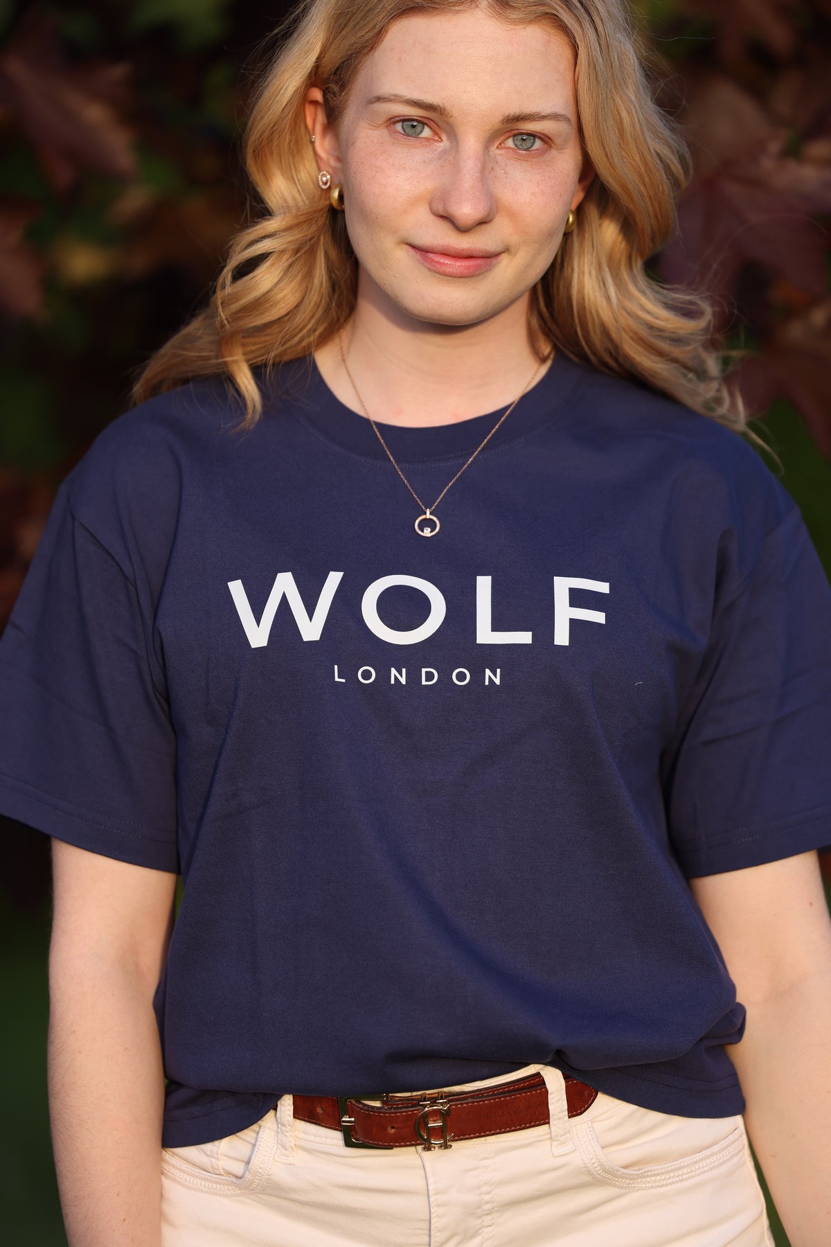 A woman wearing a navy blue t-shirt with 'WOLF LONDON' printed in white across the chest, paired with beige pants and a brown belt, set against a blurred outdoor background.